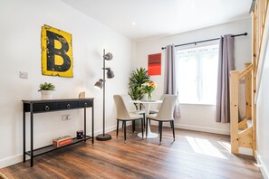 Et moderne værelse med trægulv, et skrivebord med skuffer, et rundt bord med stole, et stort bogstav B på væggen, en plante og et vindue med gardiner.