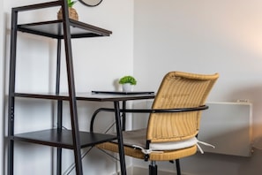 Ein Zimmer mit einem schwarzen Metallregal, einem Korbstuhl, einer kleinen Topfpflanze und einer Heizung.