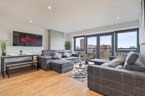 A modern living room with a large flat-screen TV, grey sofas, a coffee table, and a dining table with benches. - High-Rise 2BR | Balcony & City Views | Sleeps 8 (Leeds)