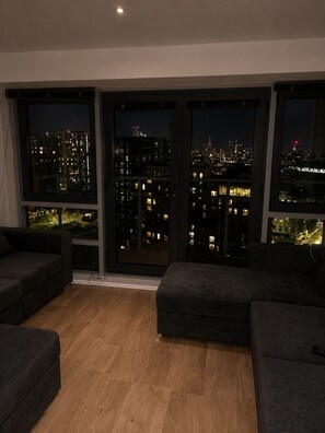 A modern living room with large windows, a dark sofa, and a wooden floor. - High-Rise 2BR | Balcony & City Views | Sleeps 8 (Leeds)