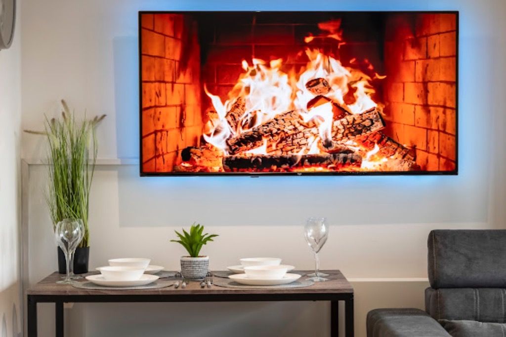 A room with a flat-screen TV displaying a fireplace, a dining table with plates and glasses, and a potted plant.