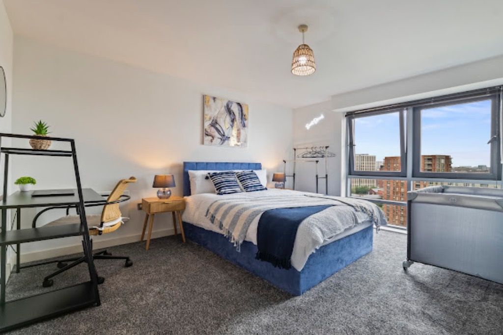 Une chambre à coucher comprenant un lit, un bureau, une chaise, une lampe, une étagère avec une plante, une fenêtre donnant sur des immeubles et un luminaire au plafond.