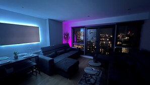 Una sala moderna con un sofá, una mesa de comedor con platos y vasos, una mesita de centro y vistas a la ciudad a través de amplias ventanas.