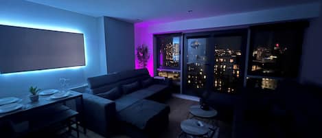 A modern living room with a sofa, a dining table with plates and glasses, a small coffee table, and a city view through large windows.