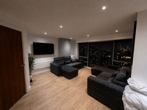 Un salon moderne doté d’un grand téléviseur à écran plat, de deux canapés sombres, d’une table basse et offrant une vue sur la ville grâce à de grandes fenêtres.