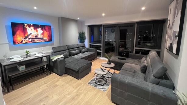 Una sala de estar moderna con televisor de pantalla plana, sofás grises, una mesa de comedor y vistas a la ciudad a través de amplios ventanales.