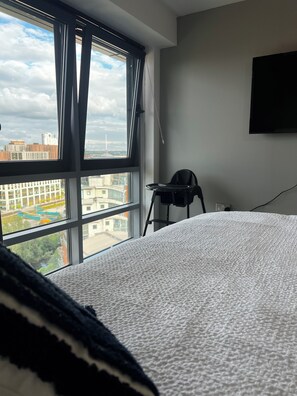 Une chambre avec une grande fenêtre, un lit recouvert d’une couverture blanche à texture, une chaise noire et un téléviseur fixé au mur.