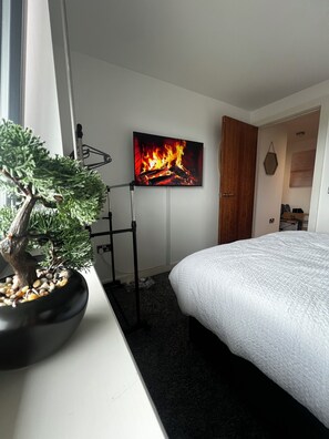 Et soveværelse med en seng, et bonsai-træ i potte, et vægmonteret TV der viser en pejs og en dør, der fører til et andet rum.