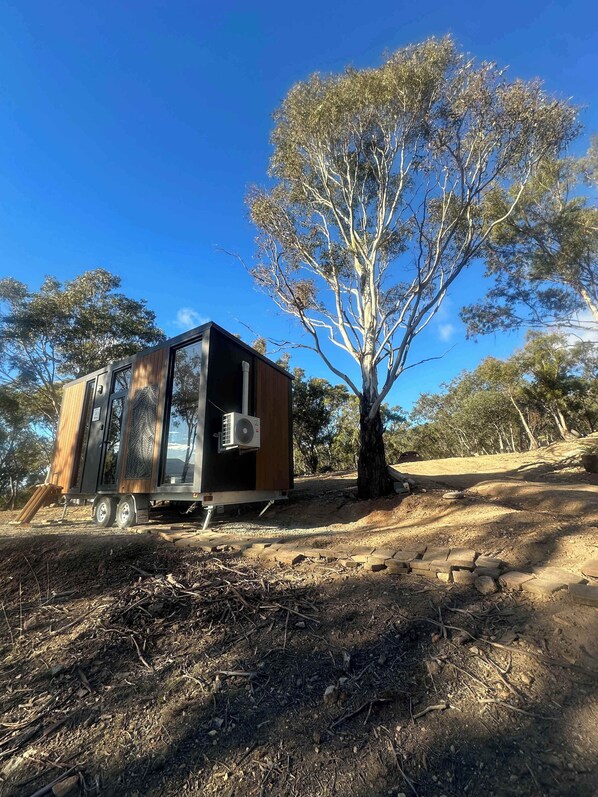 A modern, compact mobile home with a large window and a small porch area, situated on a dirt ground with trees in the background. - Black Snake Sky Box by Tiny Away (Granton)