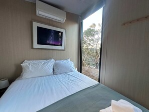 A bedroom with a bed, an air conditioner, a framed picture, and a window with a view of trees. - Black Snake Sky Box by Tiny Away (Granton)
