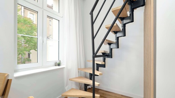 A corner room with a wooden floor, a black spiral staircase with wooden steps, and a window with white curtains. - Stylish Studio with a Mezzanine | Near the Market Square (Poznań)