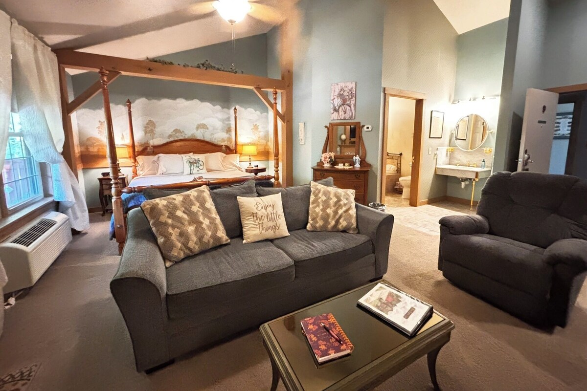 Un dormitorio con cama con dosel, una zona de estar con sofá y sillón, y un baño visible a través de una puerta abierta.