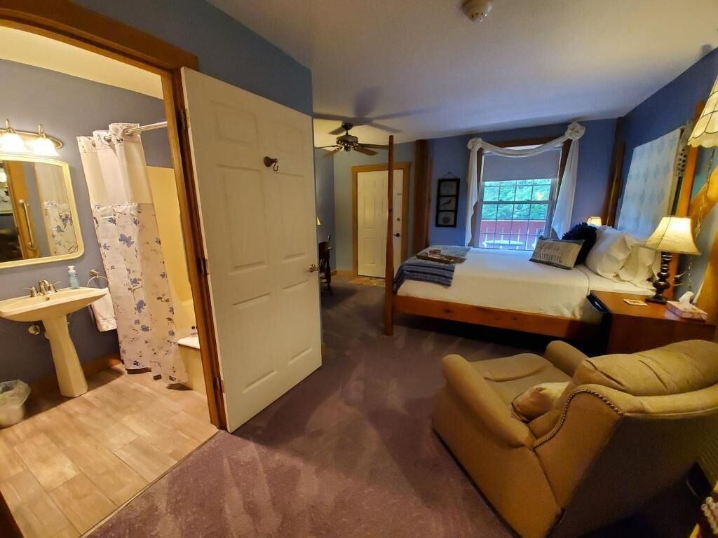 Una camera da letto con un letto, una sedia, un comodino, una lampada, una finestra e un bagno con lavandino e specchio.