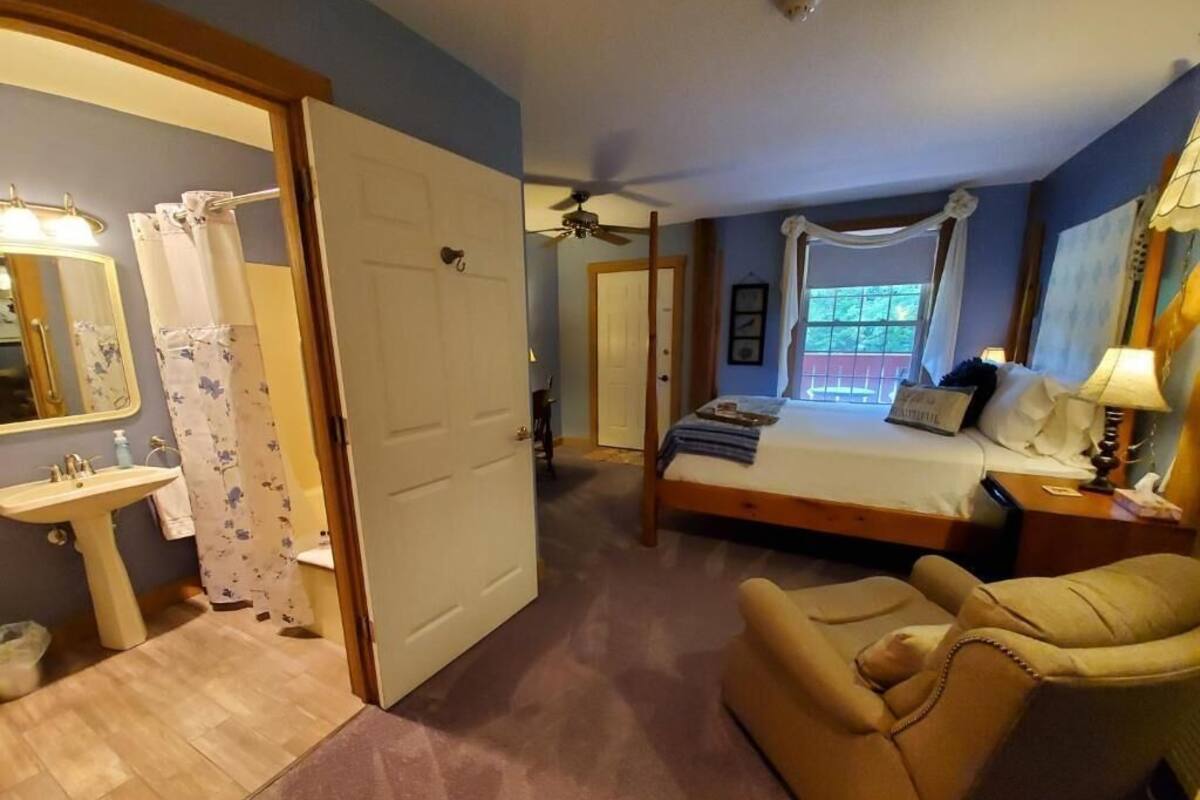 A bedroom with a bed, a chair, a bedside table, a lamp, a window, and a bathroom with a sink and mirror.