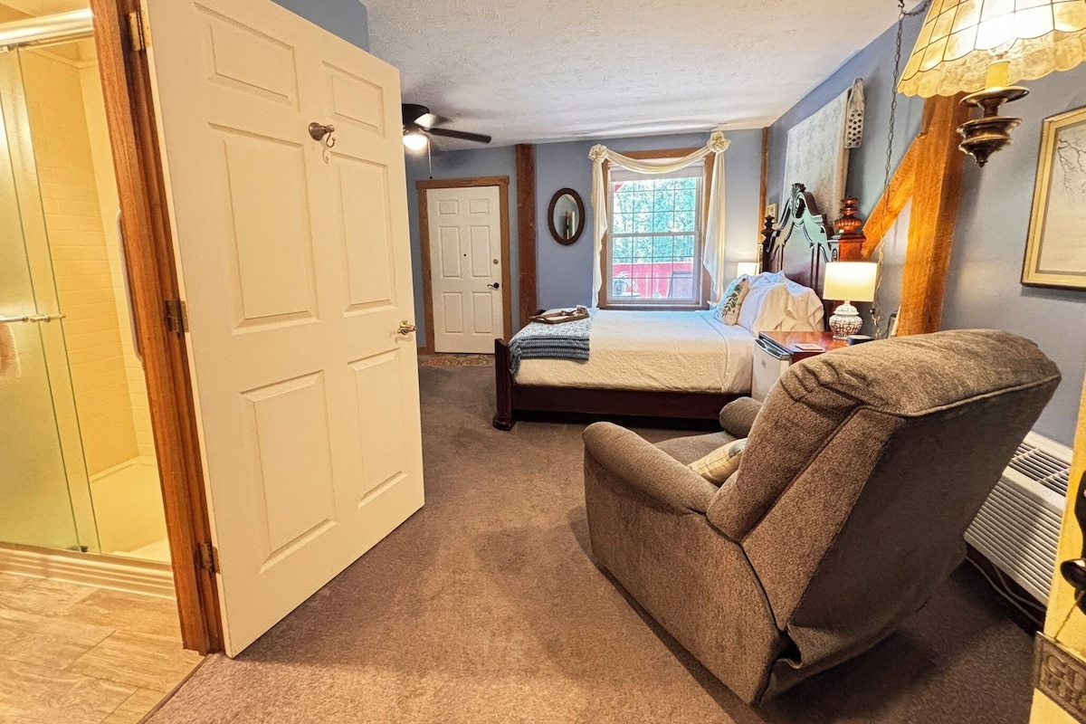 Un dormitorio con cama, una silla, ventana con cortinas, puerta y ventilador de techo.
