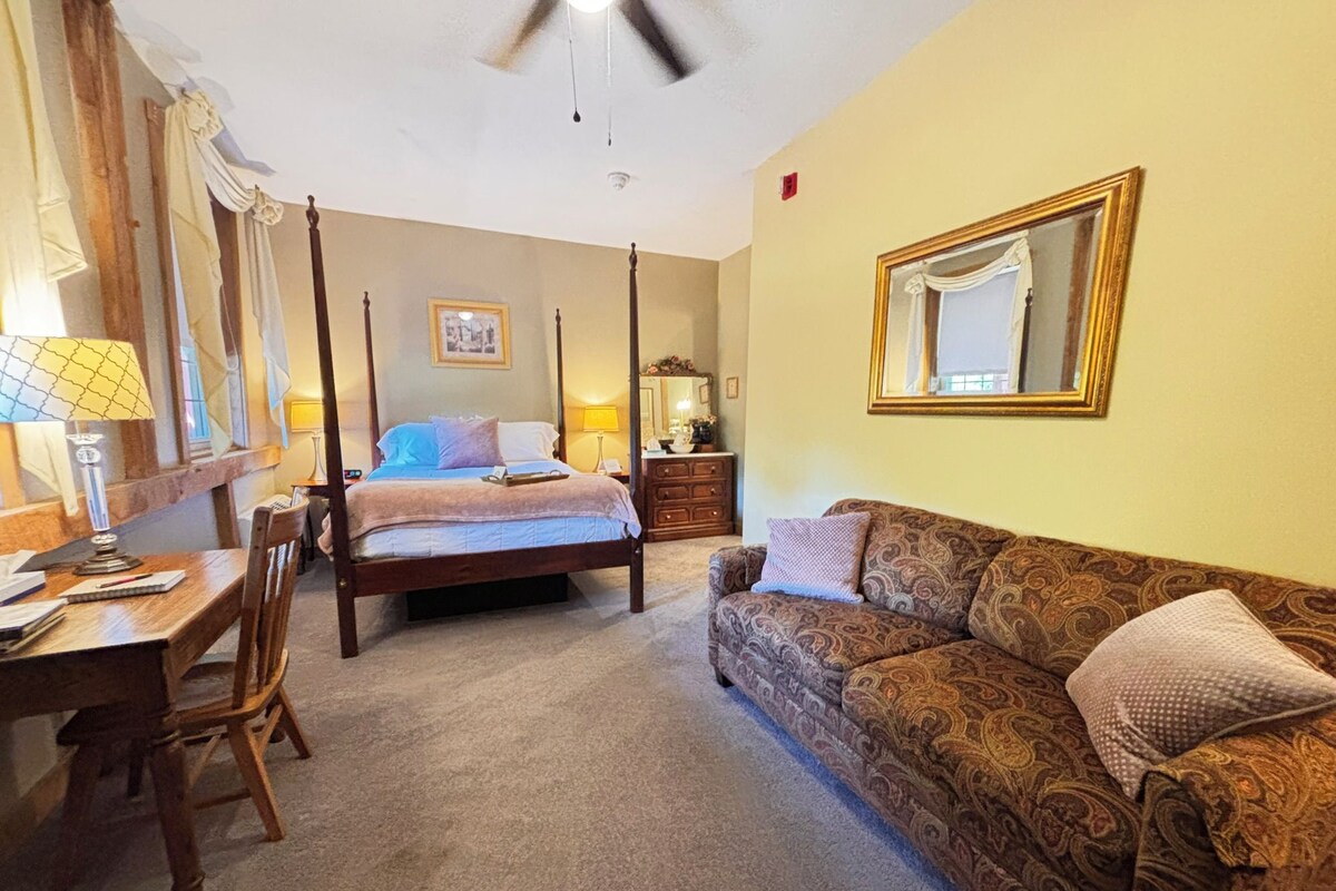 A bedroom with a four-poster bed, a wooden desk with a chair, a patterned sofa, and a large mirror on the wall.