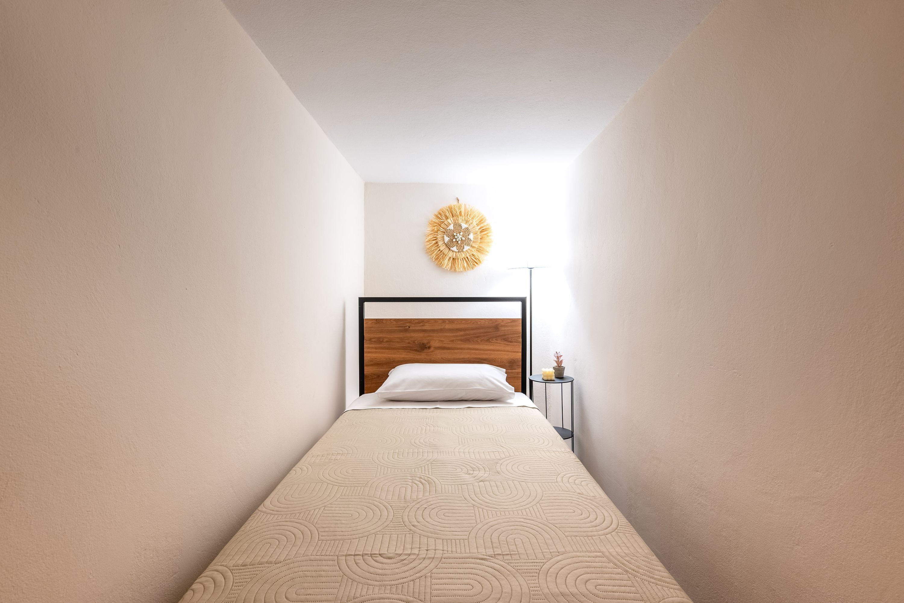 Ein schmales Zimmer mit einem Bett, einer Wandleuchte und einem kleinen Tisch mit einer Pflanze.