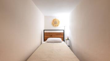 Ein schmales Zimmer mit einem Bett, einer Wandleuchte und einem kleinen Tisch mit einer Pflanze.