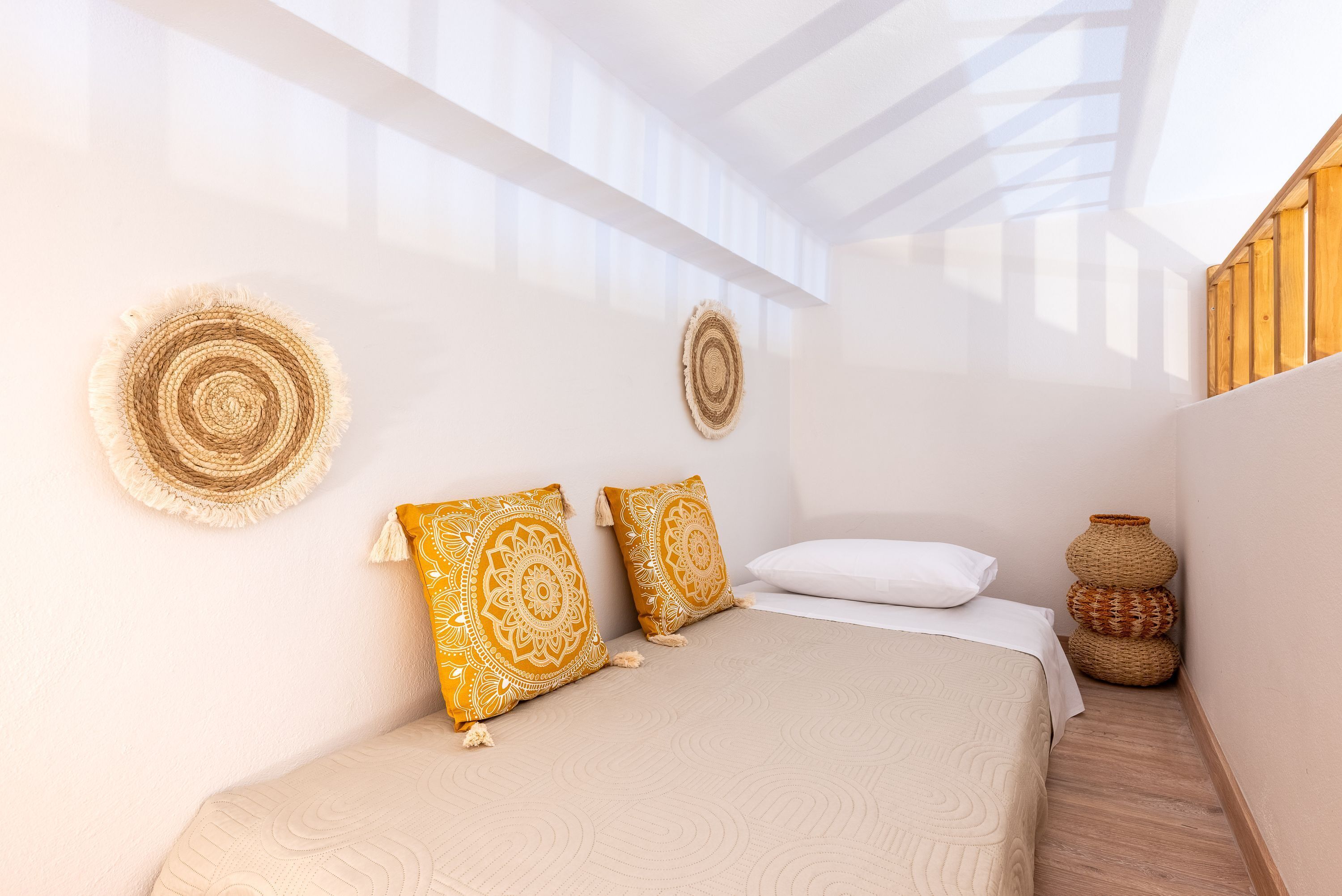 Une chambre à coucher avec un lit, deux coussins décoratifs, une tapisserie murale tissée et un panier en osier.