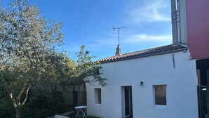 Edifici blanc amb teulada, una xemeneia i una finestra petita. Hi ha una taula i caires a l'exterior, i un arbre al fons.