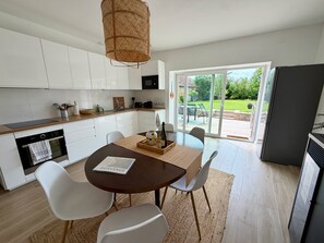 A modern kitchen with white cabinets, a dining table with chairs, a welcome mat, and a view of the outdoors through a glass door. - Vrbo Property (Périers)