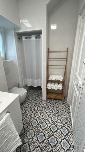 A bathroom with a toilet, a shower with a curtain, a towel rack with towels, and patterned floor tiles. - Vrbo Property (Périers)