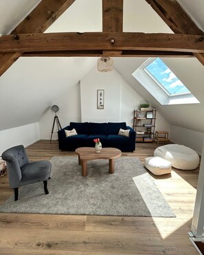 A loft-style room with a sloped ceiling, a dark blue sofa, a wooden coffee table, a grey armchair, a floor lamp, and a skylight. - Vrbo Property (Périers)
