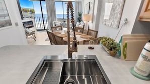 A modern kitchen with a stainless steel sink, wicker furniture, and a view of the beach.