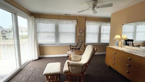 Quarto com ventoinha no teto, uma cadeira de balanço, cómoda de madeira e janelas amplas com cortinas brancas.