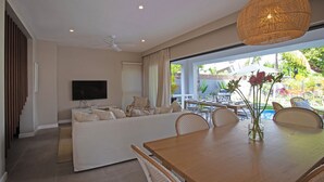 A modern living room with a sofa, TV, dining table, and wicker chairs. - Villa Andrella - Beach Haven (Pointe d'Esny)