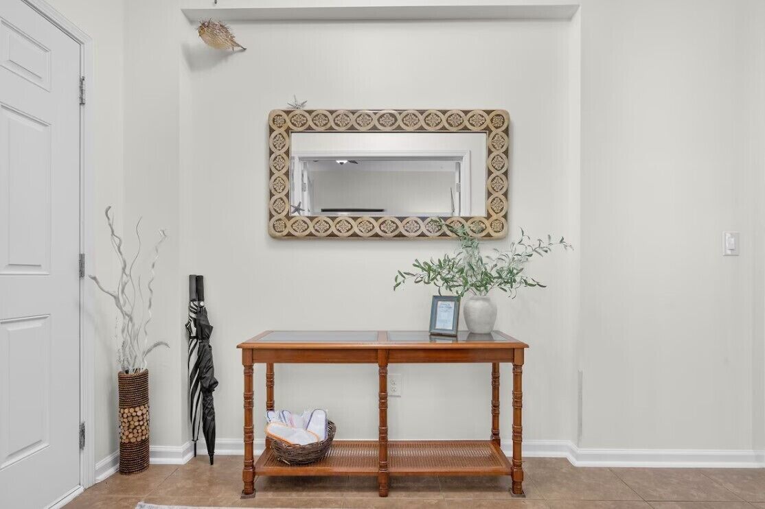 A hallway with a wooden console table, a mirror with a patterned frame, a vase with plants, and a wicker basket.