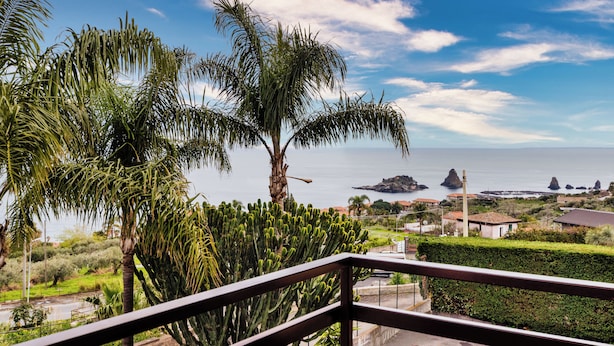 A balcony with a view of the sea, palm trees, and distant buildings. - Faraglioni Sea View Terrace by Wonderful Italy (Aci Catena)