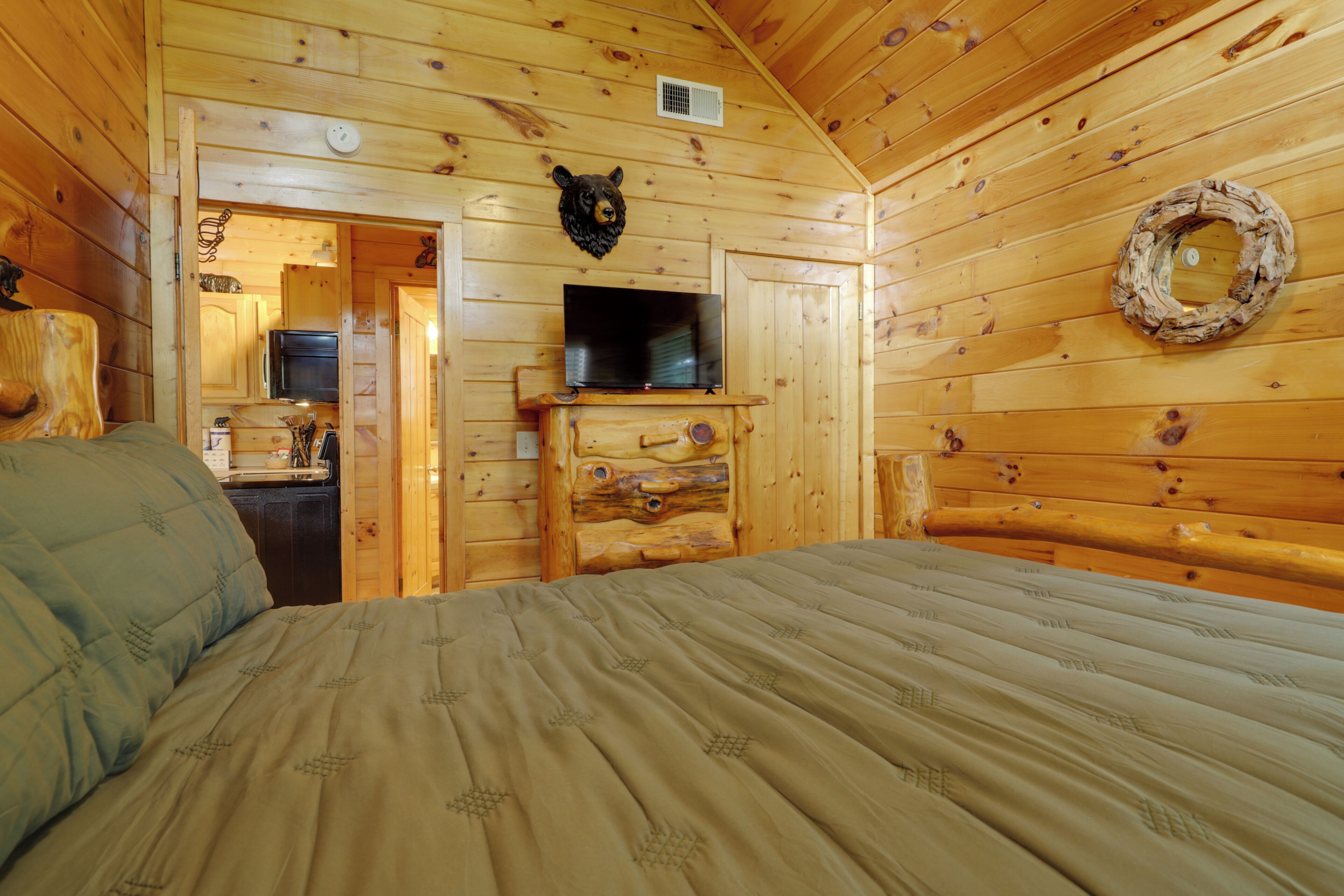 A wooden cabin interior with a bed, a television on a wooden dresser, a bear head decoration, and a wreath on the wall.