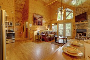 A wooden cabin interior with a kitchen, dining area, and living room, featuring a fireplace and large windows.