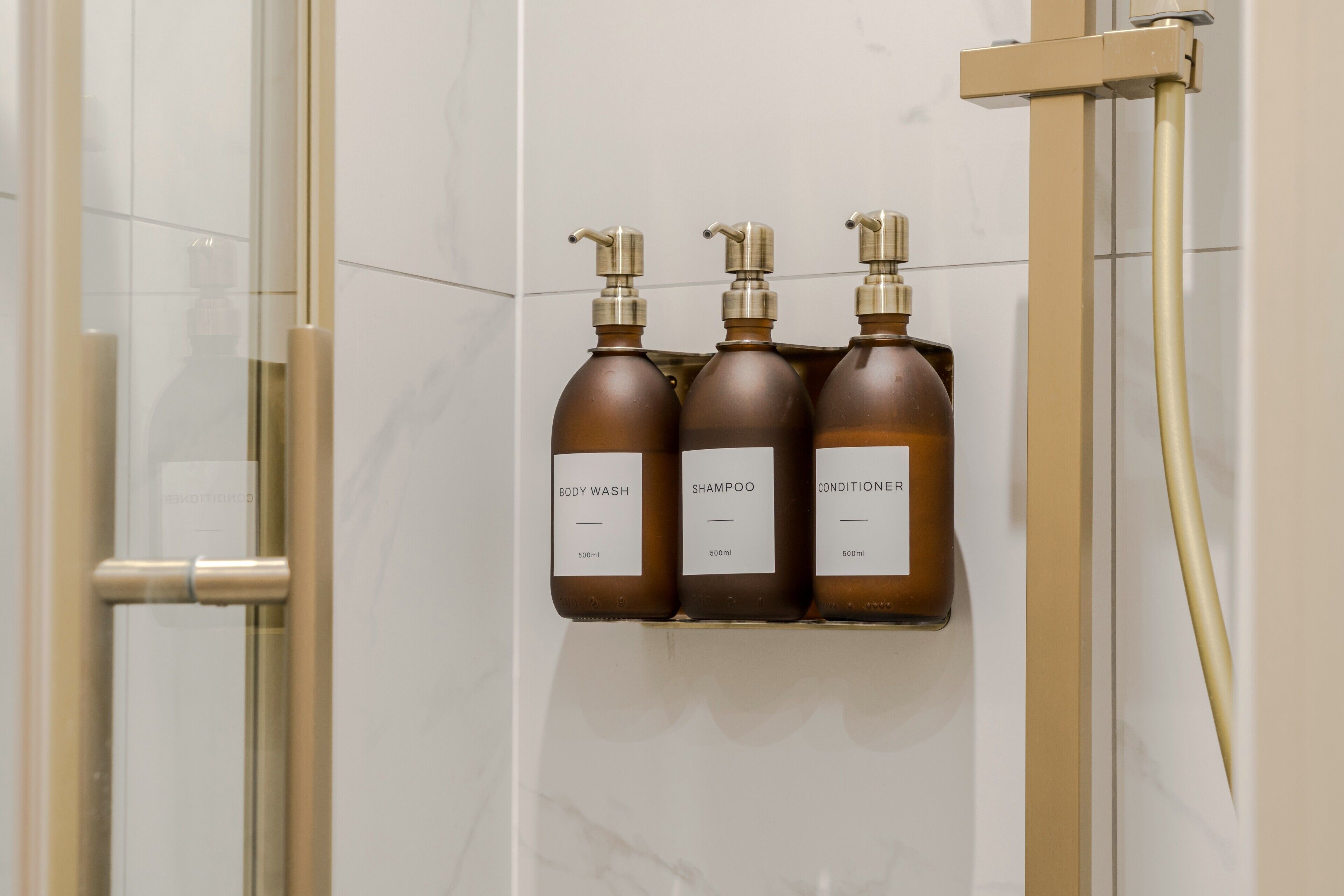 A bathroom with a glass shower enclosure, a gold showerhead, and three brown bottles of toiletries on a wall-mounted shelf.