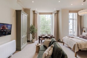 A bedroom with a bed, a green sofa, a dining table, and a potted plant. - Earl's Court - Your Apartment (London)