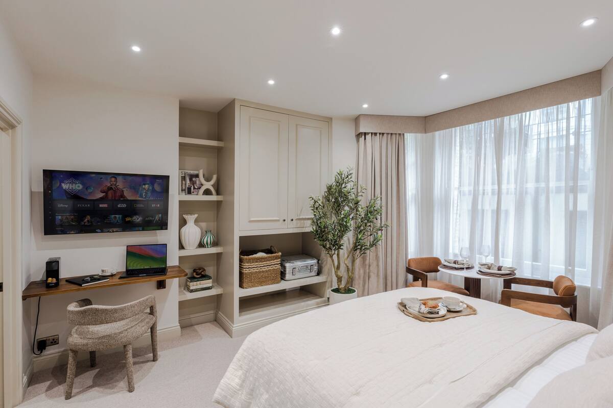 A hotel room with a bed, a desk with a laptop, a television, a plant, and a dining area with chairs and a table.