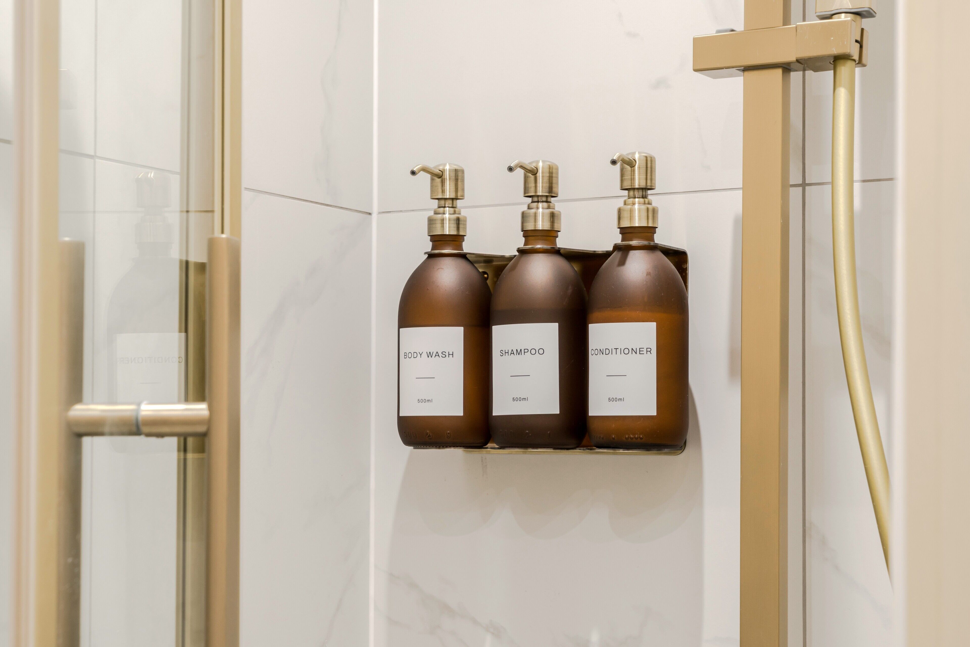 Une salle de bain avec une porte de douche en verre, une pomme de douche dorée et trois flacons de produits de toilette marron disposés sur une étagère fixée au mur.