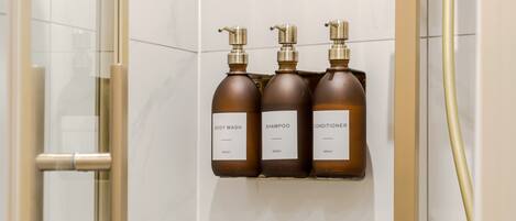 Une salle de bain avec une porte de douche en verre, une pomme de douche dorée et trois flacons de produits de toilette marron disposés sur une étagère fixée au mur.