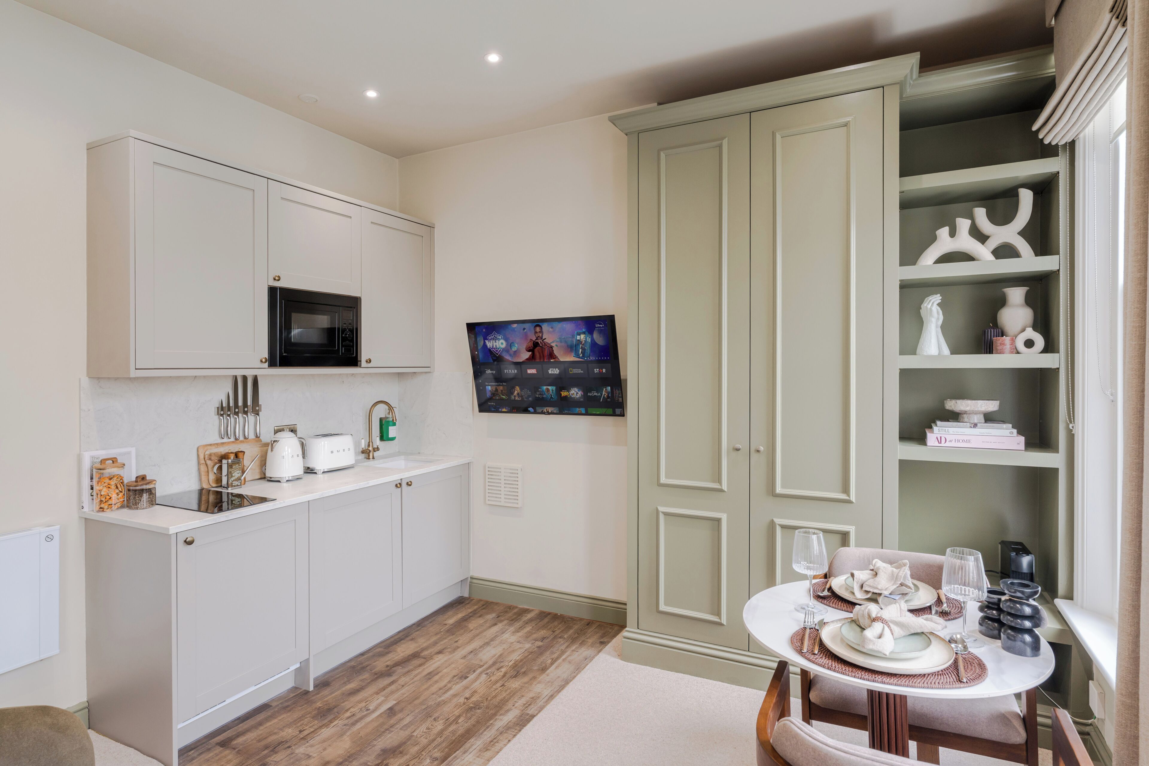 A kitchen with white cabinets, a microwave, a TV, and a dining area with a table set for two.