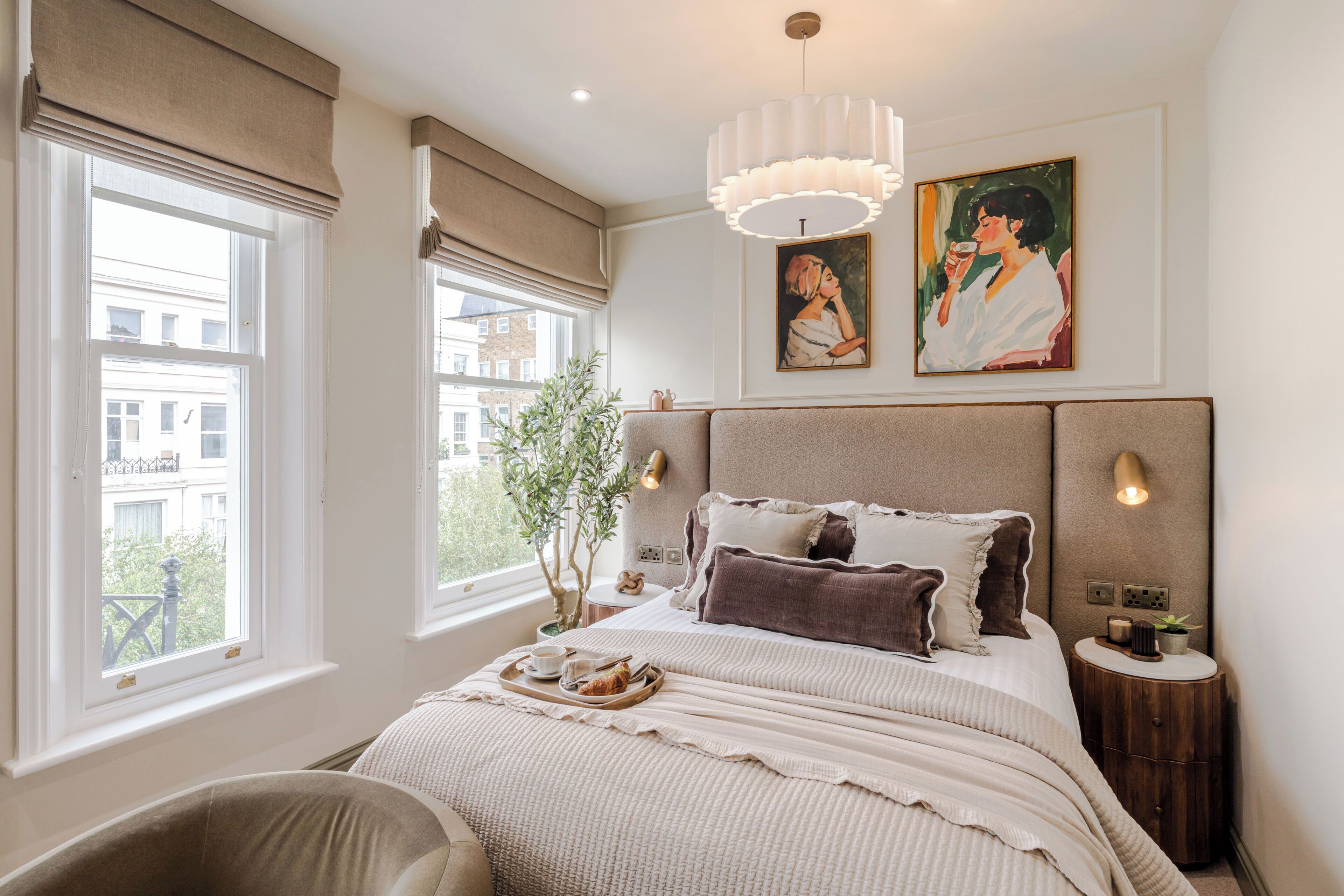 Une chambre à coucher avec un grand lit, une table de chevet, une fenêtre avec des rideaux, un luminaire suspendu et deux tableaux accrochés au mur.