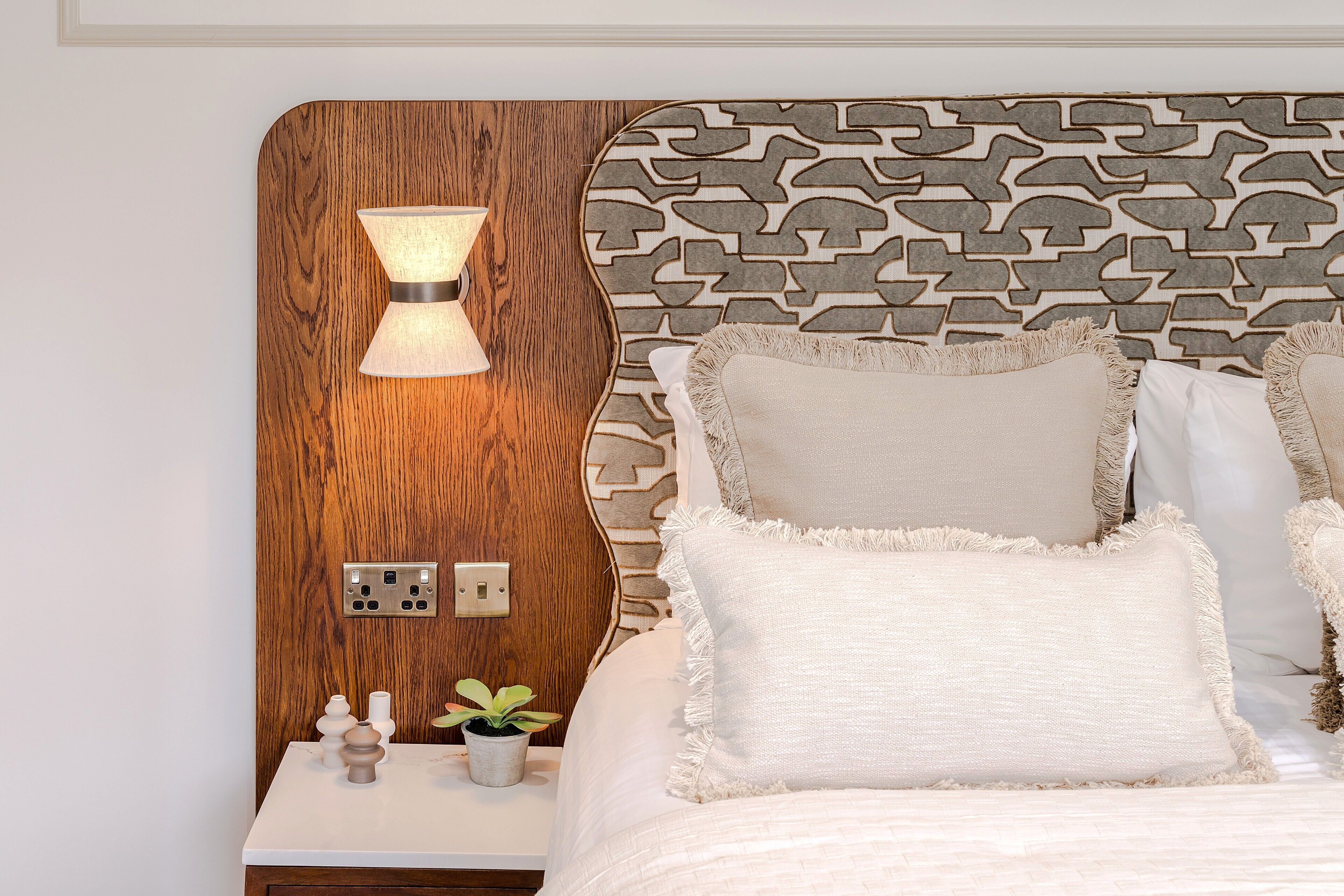 Een slaapkamer met een houten hoofdbord, een bedlamp, een nachtkastje met een plant en een bed met witte beddengoed.