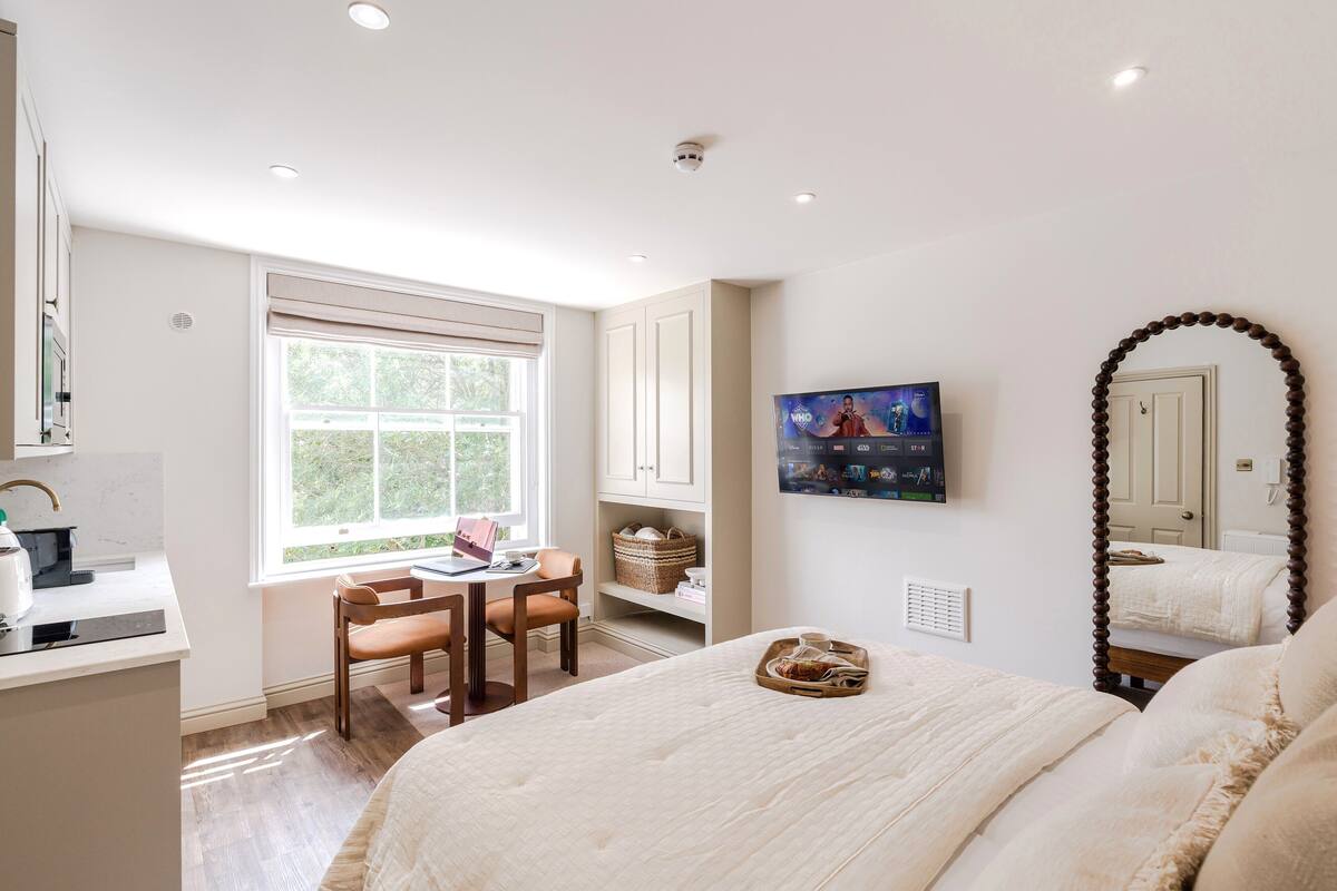A bedroom with a bed, a desk, a chair, a window, a TV, and a mirror.
