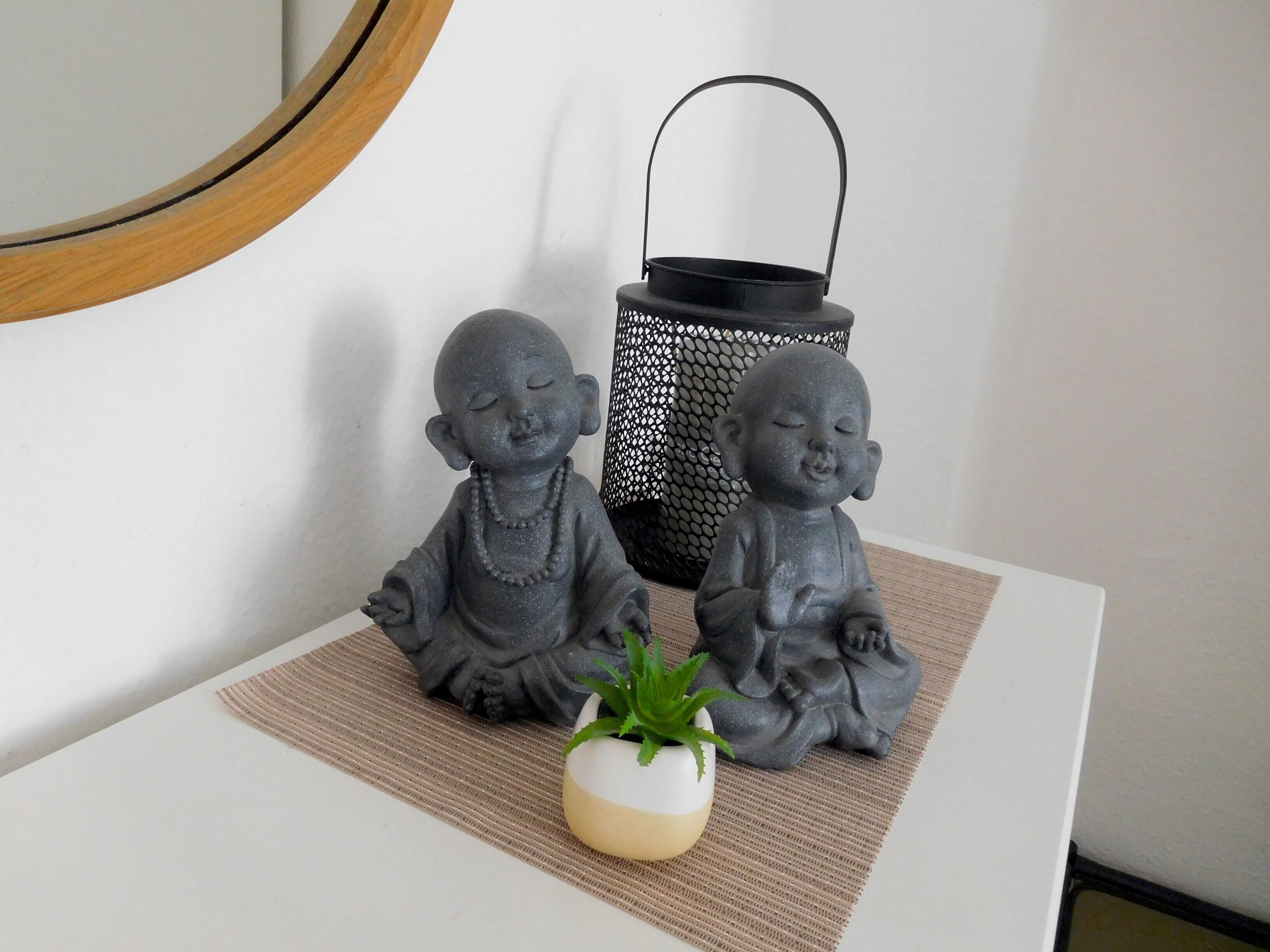 Dos estatuas de Buda sobre una mesa blanca con una pequeña planta en maceta y un farol negro.