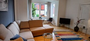 Ein Wohnzimmer mit Sofa, Glastisch, Fernseher und einem Essbereich mit Tisch und Stühlen.