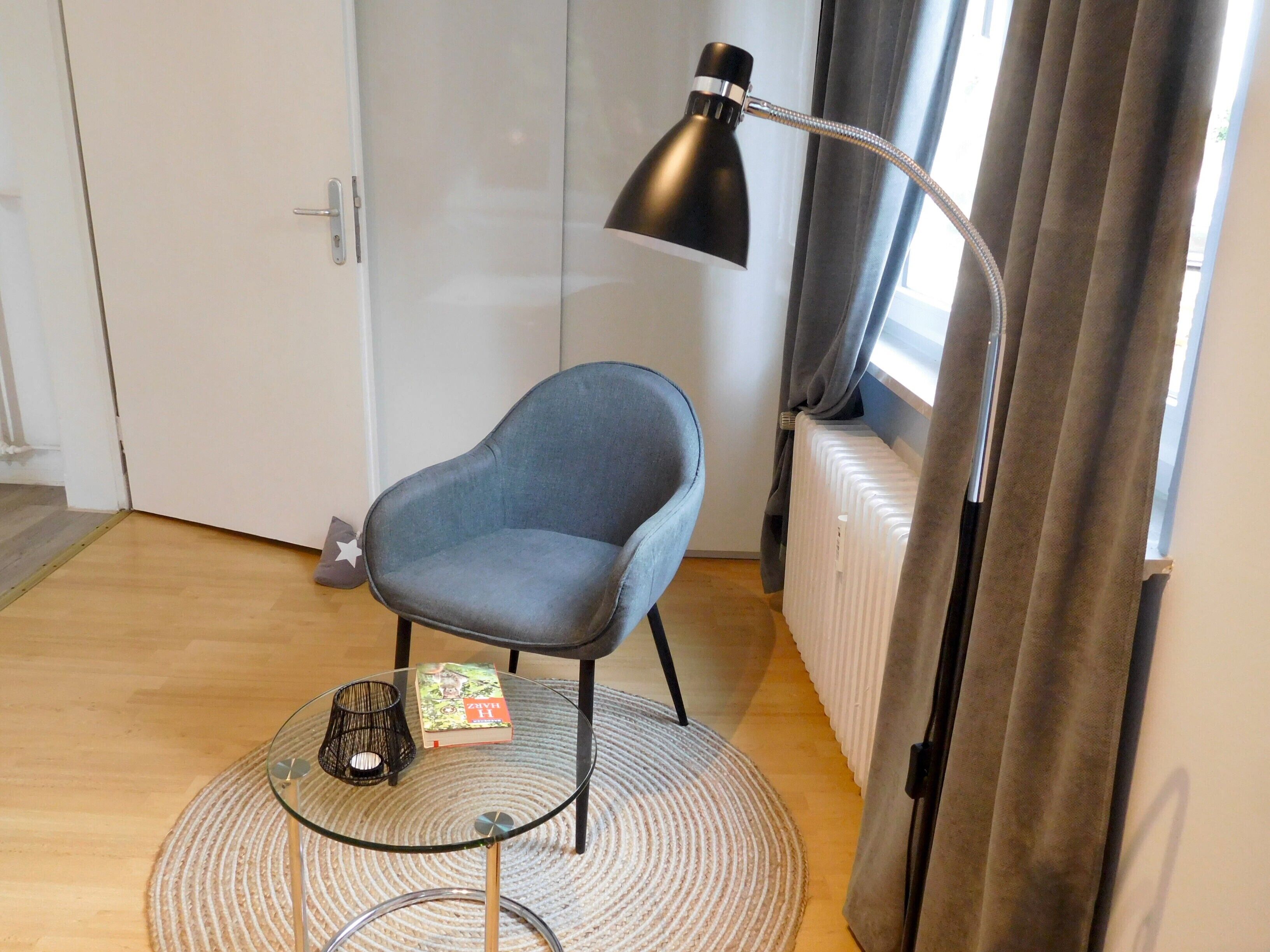 Ein Zimmer mit grauem Sessel, einem Glastisch mit Buch und Kerze, einer Stehlampe und einem Heizkörper.