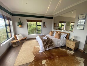 A bedroom with a large window, a bed with a cowhide rug, a wooden nightstand, and a mirror. - The Liora (hoekwil)