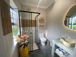 A modern bathroom with a glass shower enclosure, a toilet, a sink with a beige basin, and a mirror with a circular frame. - The Liora (hoekwil)