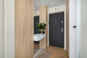Um corredor com piso de madeira, uma prateleira branca fixada na parede com plantas em vasos, uma porta escura e um interruptor de luz.