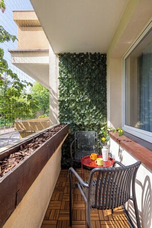 Ein Balkon mit einem Holzpflanzgefäß, einem Metallstuhl, einem kleinen Tisch mit Tablett und einem Fenster mit Blick auf Grünflächen.